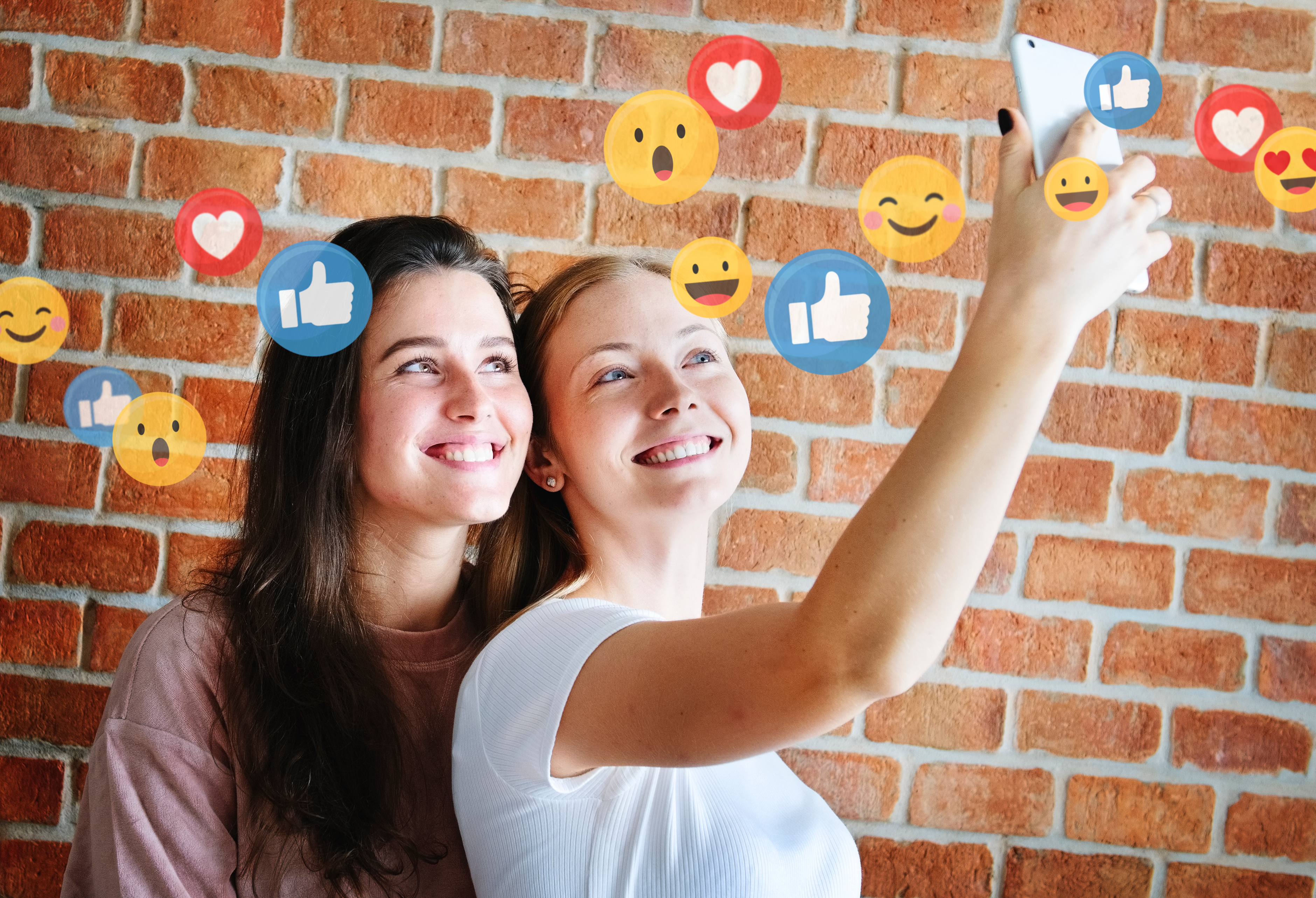 Two young women take a selfie together. Above them are social media icons such as emojis, hearts and thumbs up. A brick wall can be seen in the background.
