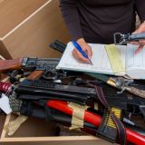 Mehrere Waffen liegen übereinander. Darüber trägt eine Person etwas in ein Buch ein. 