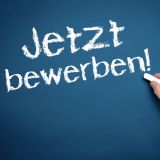 Eine Hand schreibt mit Kreide "Jetzt bewerben!" an eine Tafel