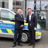 Landrat Andreas Müller schüttelt dem Leitenden Polizeidirektor Klaus Bunse die Hand und begrüßt ihn als neuen Abteilungsleiter der Polizei Siegen-Wittgenstein. Die beiden Personen stehen vor einem Polizeifahrzeug. Im Hintergrund ist der Haupteingang des Polizeigebäudes in Siegen-Weidenau zu sehen.