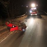 Auf einer dunklen Landstraße liegt ein roter Opel Astra am linken Straßenrand auf dem Fahrzeugdach. Im Hintergrund leuchtet ein Einsatzfahrzeug der Feuerwehr die Unfallstelle aus.