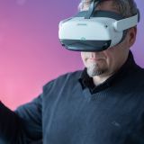 Ein Mann trägt eine VR-Brille und hält einen Controller in der Hand. Im Hintergrund ist ein Bildschirm mit einem rosa Farbverlauf zu sehen.