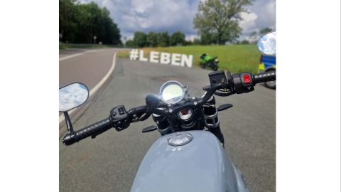 Man blickt über den Tank und Lenker eines silberfarbenen Motorrades. Im Hintergrund sind die Buchstanden Hashtag LEBEN in einer Haltebucht neben einer Landstraße aufgebaut. Rechts daneben befindet sich ein abgestelltes grünes Motorrad.