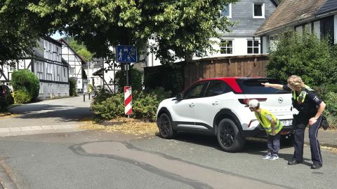Eine Polizeibeamtin hilft einem Kind beim sicheren Überqueren der Straße hinter einem am Straßenrand geparkten Pkw