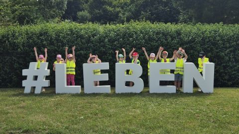 Man sieht die Kampagnenbuchstaben Hashtag Leben draußen im Freien vor einer grünen Hecke. Hinter den Buchstaben stehen Kinder mit Leuchtwesten, die die Arme hochstrecken.