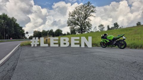 Neben einer Landstraße stehen die Buchstaben #LEBEN auf einem asphaltierten Parkstreifen. Rechts neben den Buchstaben steht ein grünes Motorrad abgestellt.