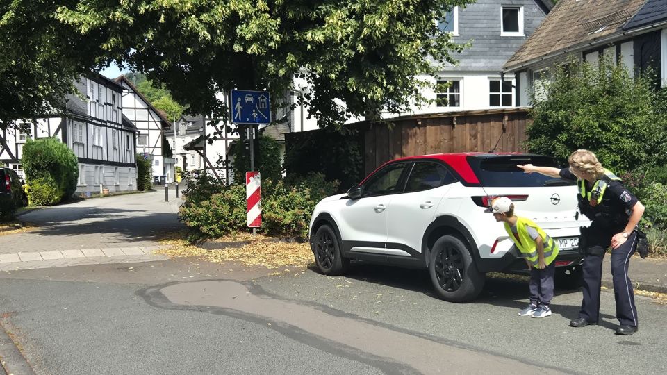 Eine Polizeibeamtin hilft einem Kind beim sicheren Überqueren der Straße hinter einem am Straßenrand geparkten Pkw