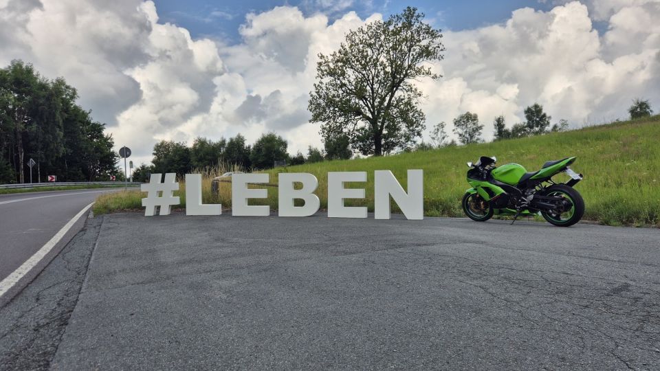 Neben einer Landstraße stehen die Buchstaben #LEBEN auf einem asphaltierten Parkstreifen. Rechts neben den Buchstaben steht ein grünes Motorrad abgestellt.
